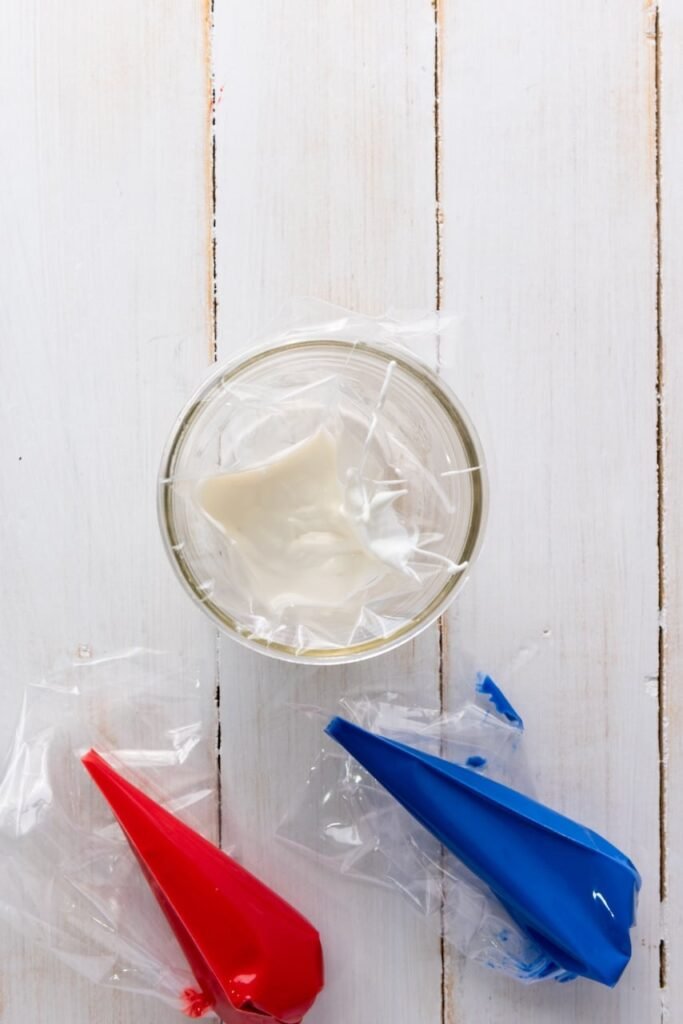 A bowl of melted white candy melts surrounded by red and blue candy melts in piping bags on a white wooden surface, showing how to prepare for drizzling popcorn.