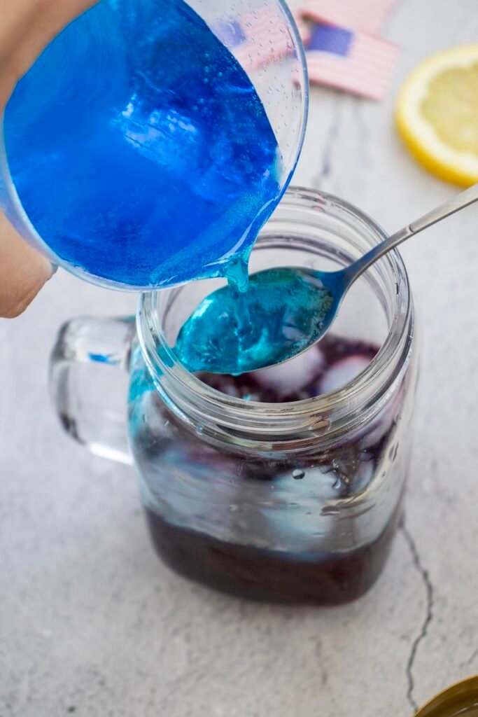 Close-up of blue sports drink being slowly poured over a spoon into a glass mug with ice to form the top blue layer of a 4th of july drink.