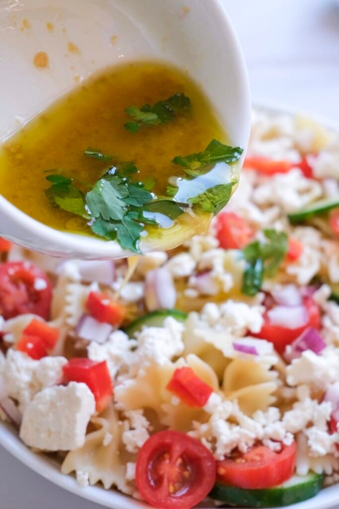 Close-up of homemade vinaigrette dressing being poured over bowtie pasta salad with cherry tomatoes, cucumber, red onion, and crumbled feta cheese in a white bowl.