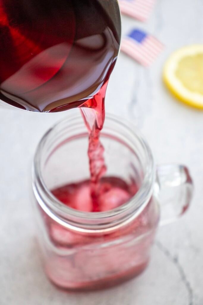 Close-up of cranberry juice being poured into a glass mug filled with ice to create the red layer of a 4th of july mocktail.