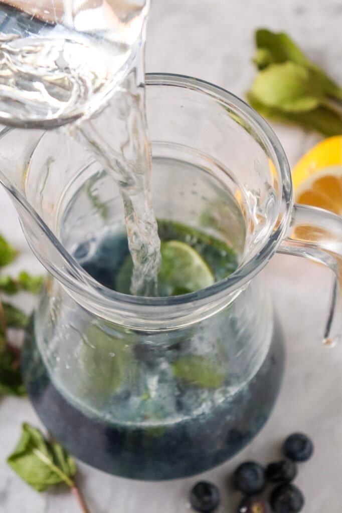 Sparkling water being poured into a glass pitcher with blueberries and mint to finish.