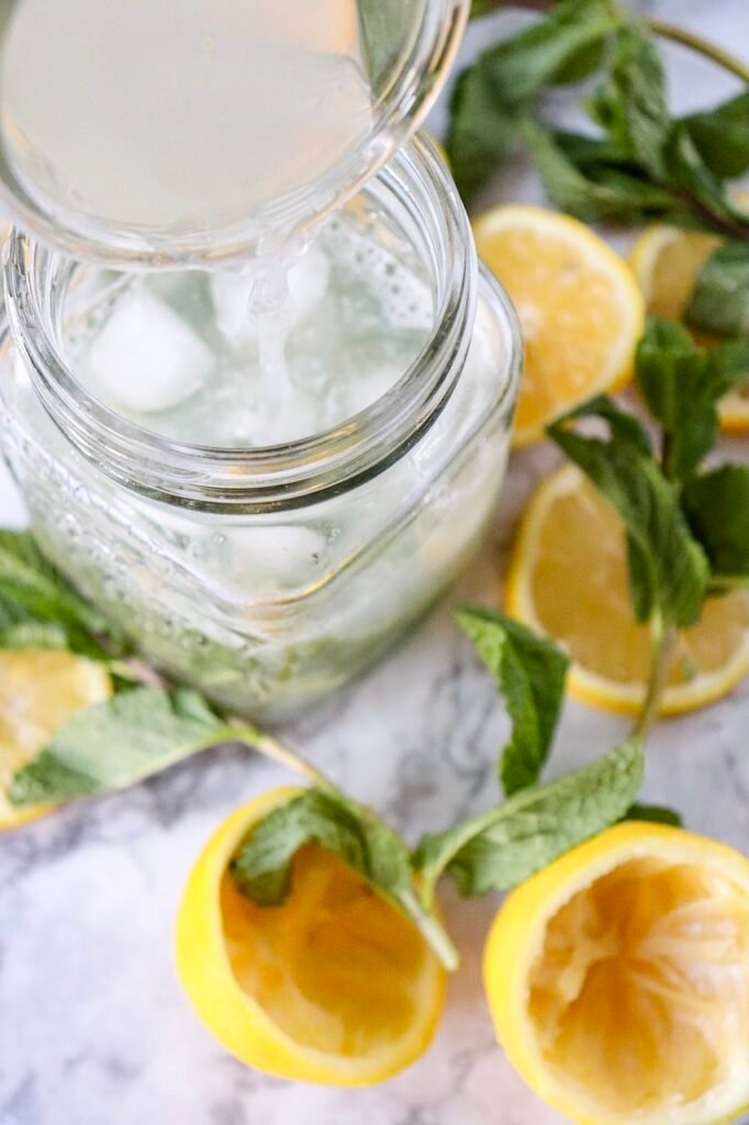 Close-up of sparkling water being poured into a glass jar with ice, lemon juice, and mint, surrounded by lemon halves and fresh mint for a virgin citrus drink
