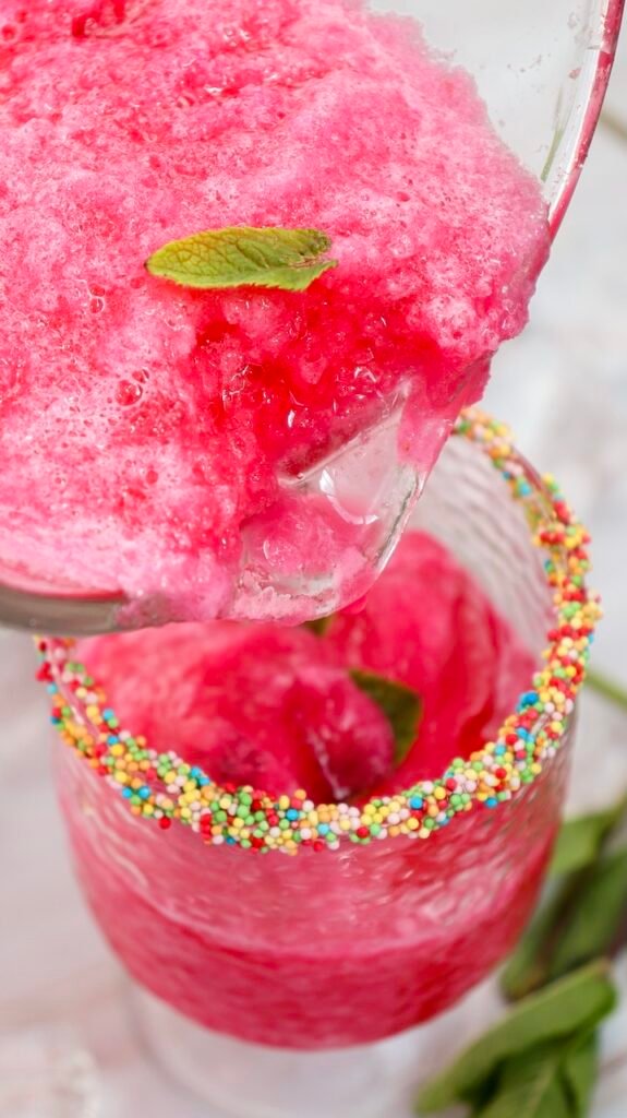 Close-up of summer drink being poured into a glass with a colorful sprinkle rim and mint garnish, showing the final serving step