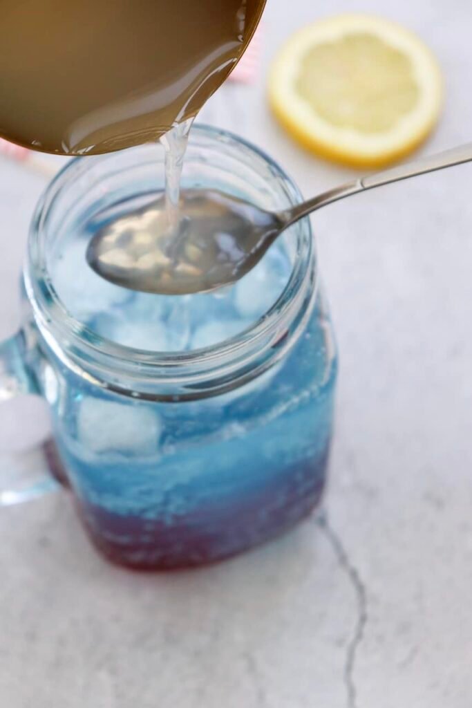 Close-up of lemonade being poured over a spoon into a glass mug with ice to form the white middle layer of a patriotic punch.