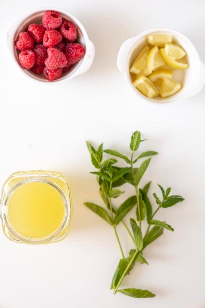 Overhead view of fresh raspberries, sliced lemon, mint sprigs, and a glass of juice arranged on a white surface for making a raspberry mojito mocktail.