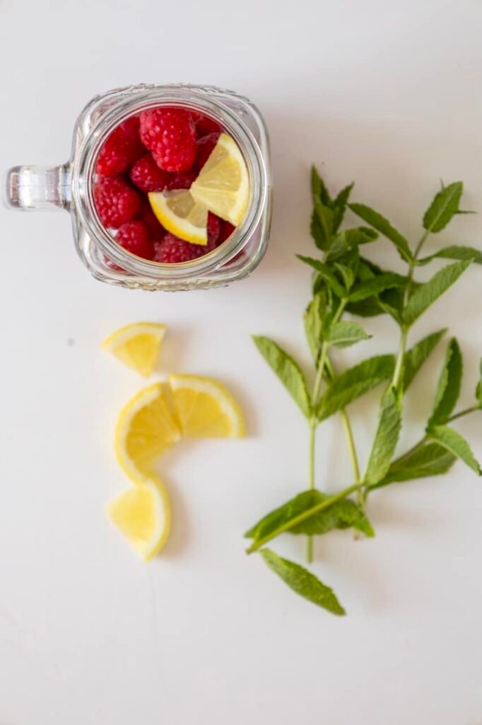 Overhead view of raspberries and lemon slices inside a glass jar with fresh mint beside it, showing ingredients for a raspberry mojito mocktail.