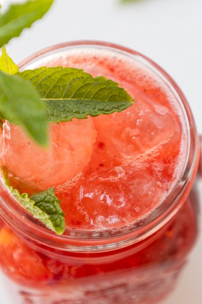 Close-up of a finished drink in a glass jar with ice, fresh mint leaves, and a bubbly raspberry mixture.
