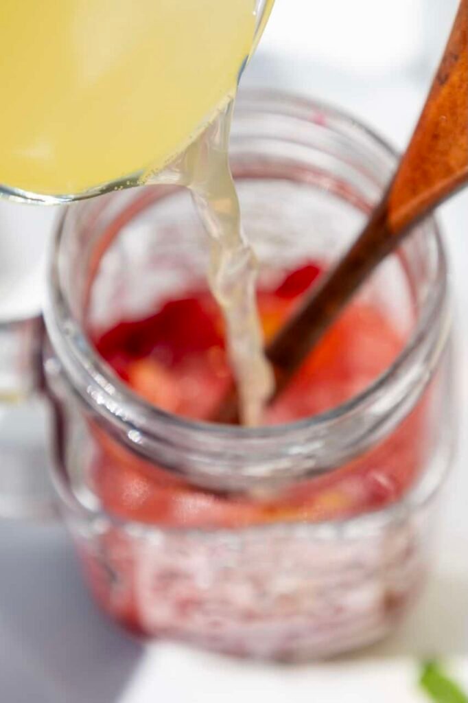 Sparkling water being poured into a jar with muddled raspberries and lemon while a wooden spoon stirs the mixture.