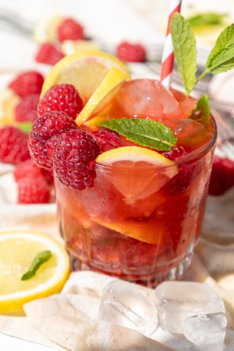 Glass of raspberry mojito mocktail filled with ice, fresh raspberries, lemon slices, and mint leaves, styled with extra fruit in the background.
