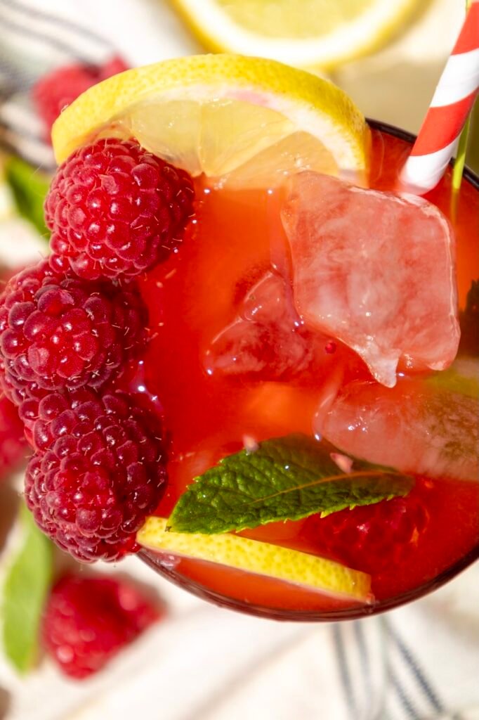 Close-up of a raspberry mojito mocktail showing fresh raspberries, lemon slices, mint leaves, and ice in a glass with a straw.