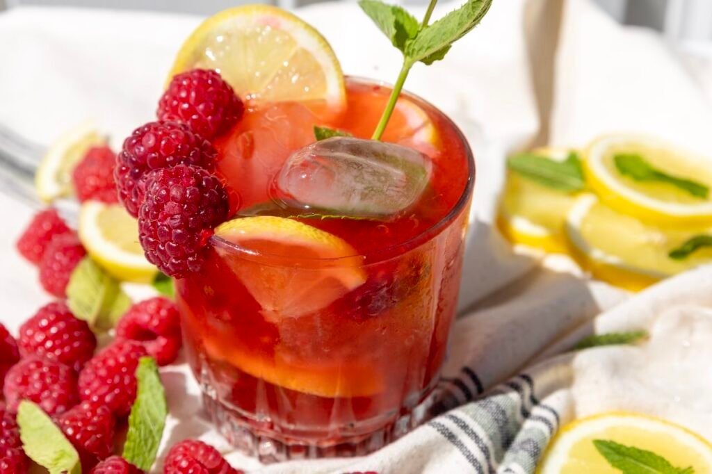 Glass of raspberry mojito mocktail with ice, fresh raspberries, lemon slices, and mint garnish, styled with extra fruit and ice in the background.