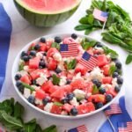 Bowl of watermelon feta salad with blueberries, mint, and red onion, decorated with small American flags and a halved watermelon in the background.