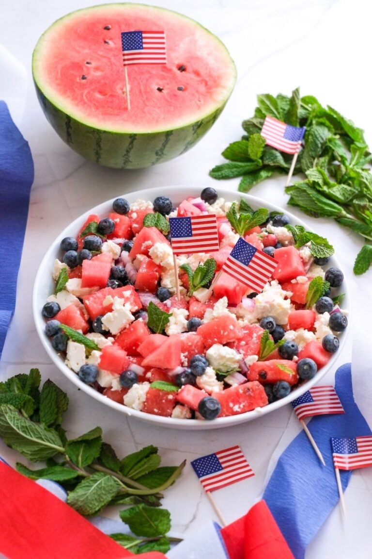 Bowl of watermelon feta salad with blueberries, mint, and red onion, decorated with small American flags and a halved watermelon in the background.