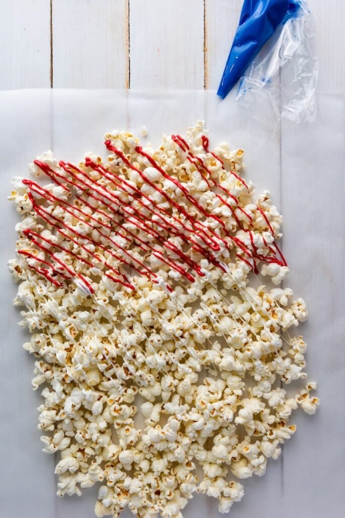 Popcorn spread on parchment paper with red and white candy melts drizzled over the top, with a blue piping bag nearby, showing a step in making festive 4th of July popcorn.