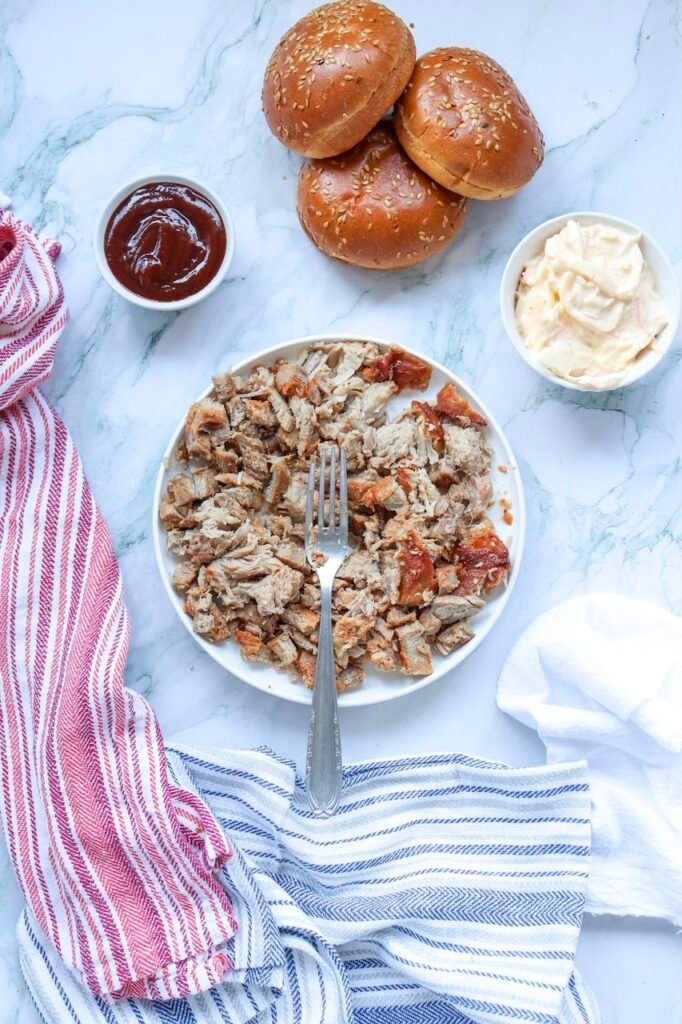 Plate of shredded cooked pork with a fork, surrounded by sandwich buns, BBQ sauce, and coleslaw, showing the prepared filling for pulled pork sandwiches.
