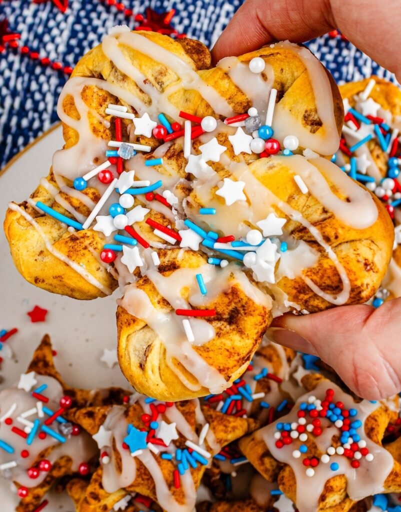Hand pulling apart a soft star-shaped cinnamon roll topped with icing and red, white, and blue sprinkles on a plate.