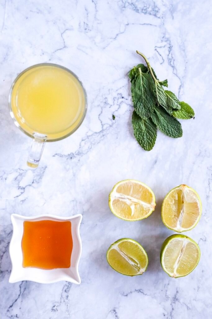 Overhead view of fresh ingredients for a virgin mojito mocktail including halved limes, mint leaves, agave syrup in a small dish, and a cup of lime juice on a marble surface.