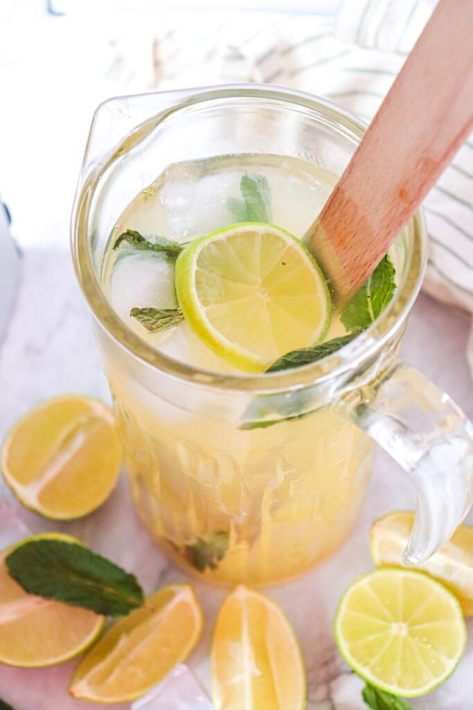 Glass pitcher with lime slices, mint leaves, and ice being stirred with a wooden spoon on a marble surface.