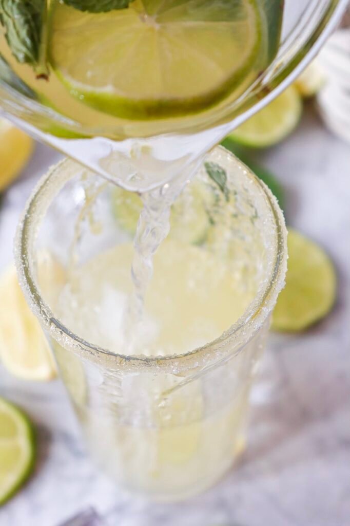 Virgin mojito mocktail being poured from a pitcher into a sugar-rimmed glass filled with ice, lime slices, and mint.