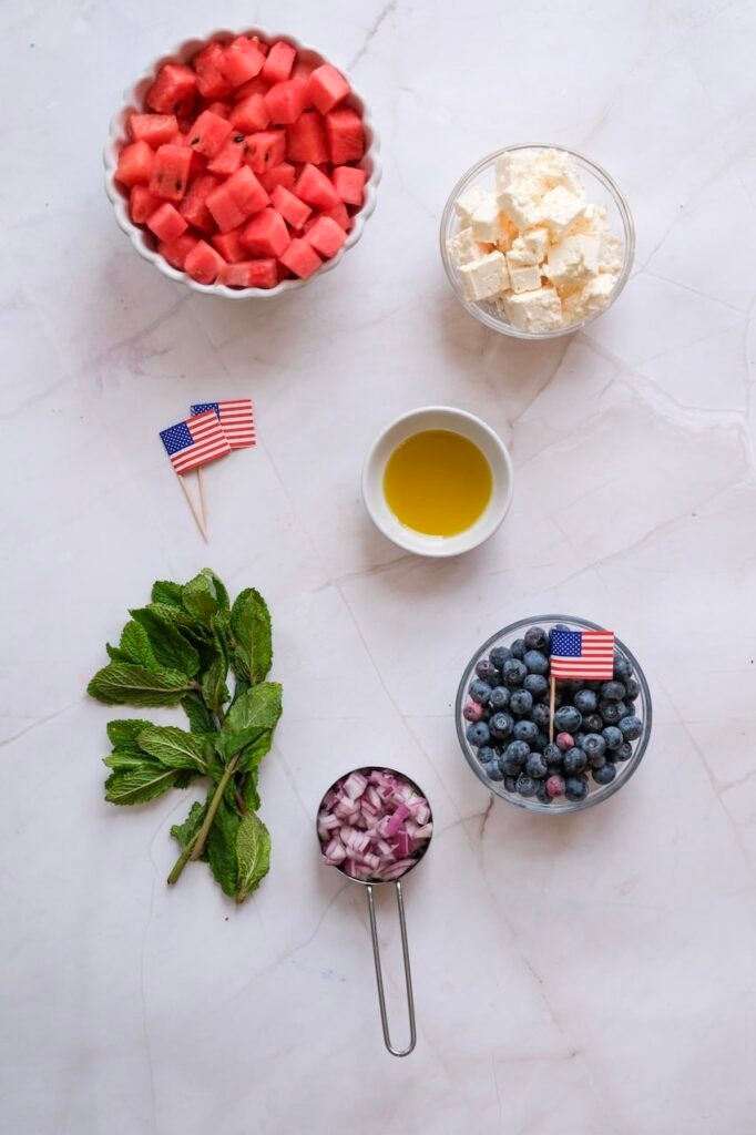 Ingredients for watermelon feta salad arranged on a light surface, including diced watermelon, blueberries, crumbled feta cheese, fresh mint, red onion, and olive oil in small bowls.