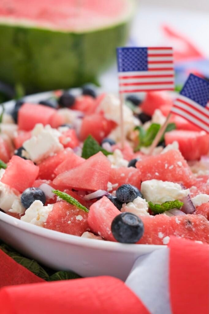 Close-up side view of watermelon feta salad with blueberries, mint, and small American flags in a serving bowl.