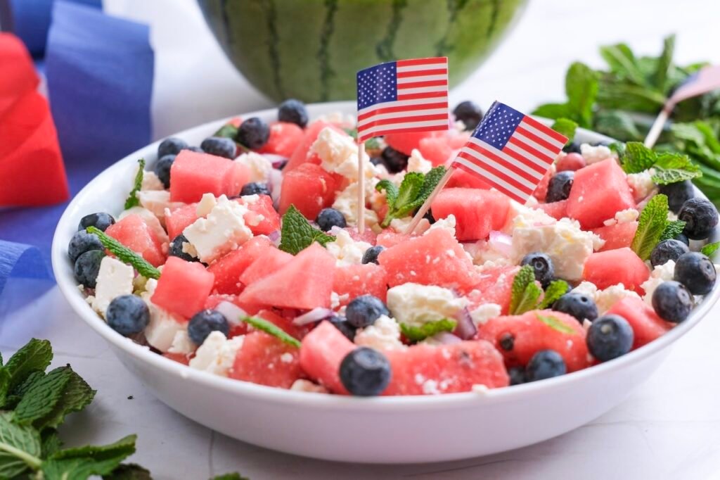 Bowl of watermelon feta salad with blueberries, mint, and small American flags, surrounded by red, white, and blue decorations.