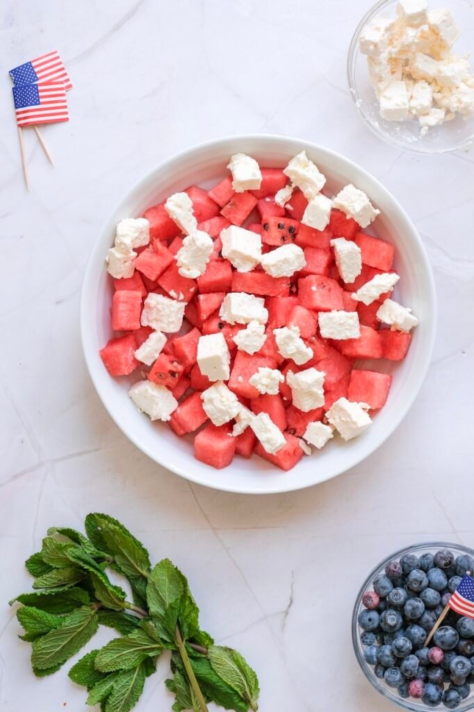 Bowl of diced fruit topped with crumbled cheese, with blueberries and fresh mint nearby for making side dish.