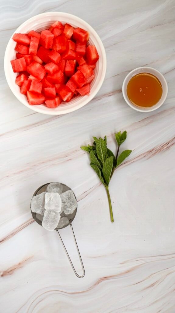 Top view of cubed watermelon in a bowl with ice cubes, honey in a small dish, and fresh mint on a marble surface, showing ingredients for a watermelon slushie