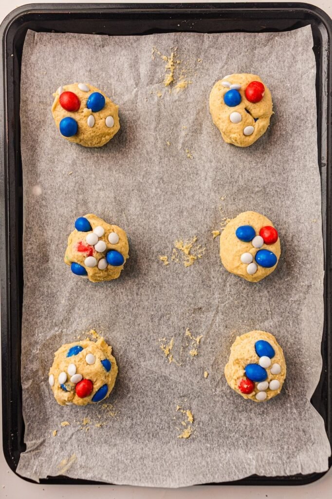 Scooped cookie dough balls topped with red, white, and blue M&Ms arranged on a parchment-lined baking tray before baking. The thick homemade cookie dough portions are evenly spaced to create soft and chewy patriotic cookies with bakery-style texture and colorful candy tops.