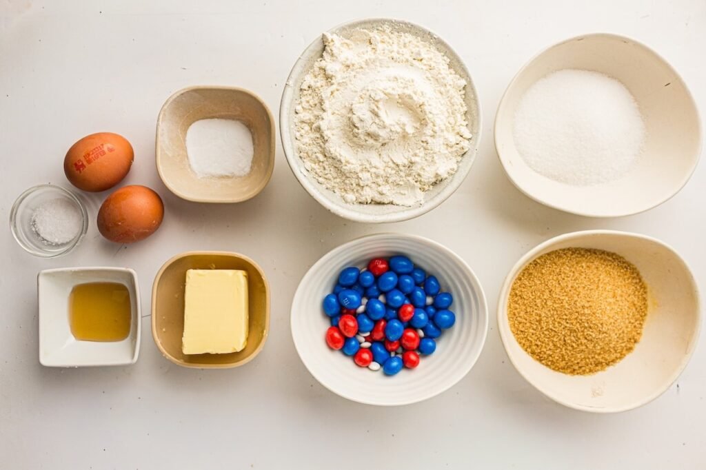 Overhead view of ingredients for homemade 4th of July cookies arranged in ceramic bowls on a white surface, including flour, white sugar, brown sugar, butter, eggs, vanilla extract, baking soda, salt, and red, white, and blue M&Ms.