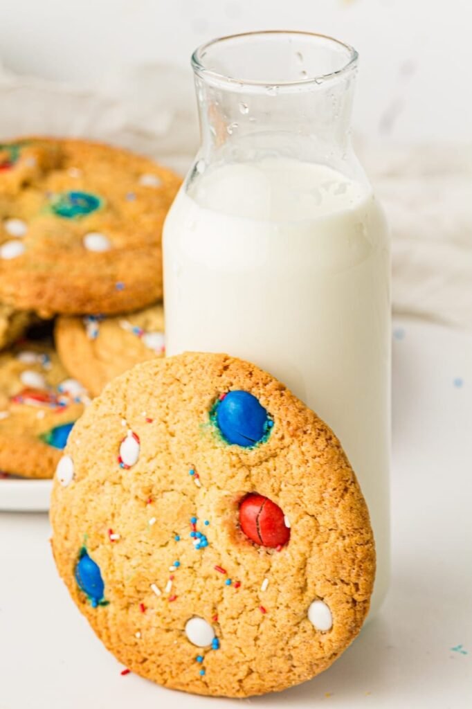 Large soft and chewy 4th of July cookie with red, white, and blue M&Ms leaning against a glass bottle of milk with more patriotic cookies blurred in the background. The homemade patriotic cookies have golden brown edges, bakery-style texture, and festive sprinkles perfect for summer holiday desserts and Independence Day parties.