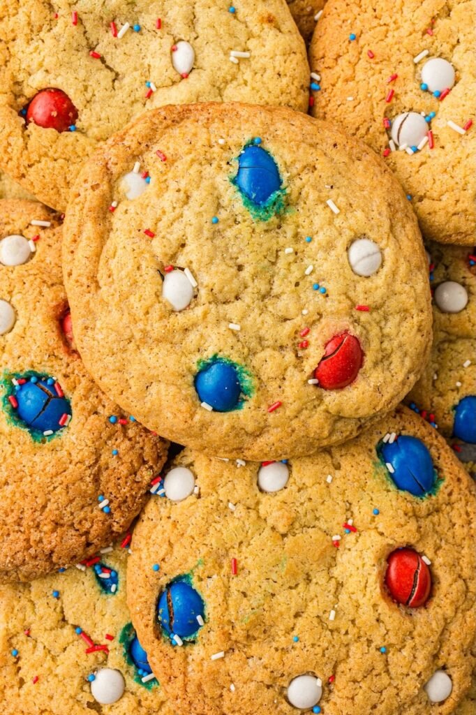 Close-up overhead view of soft and chewy 4th of July cookies layered together with red, white, and blue M&Ms and patriotic sprinkles baked into the tops. The homemade patriotic cookies have golden brown edges, thick soft centers, and bakery-style texture perfect for summer holiday dessert tables and Independence Day celebrations.