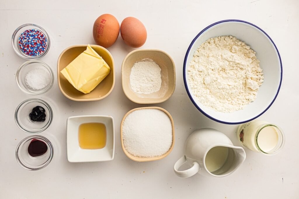 Ingredients for homemade 4th of July cupcakes arranged on a white table, including flour, sugar, butter, eggs, milk, vanilla, food coloring, sprinkles, and baking powder.