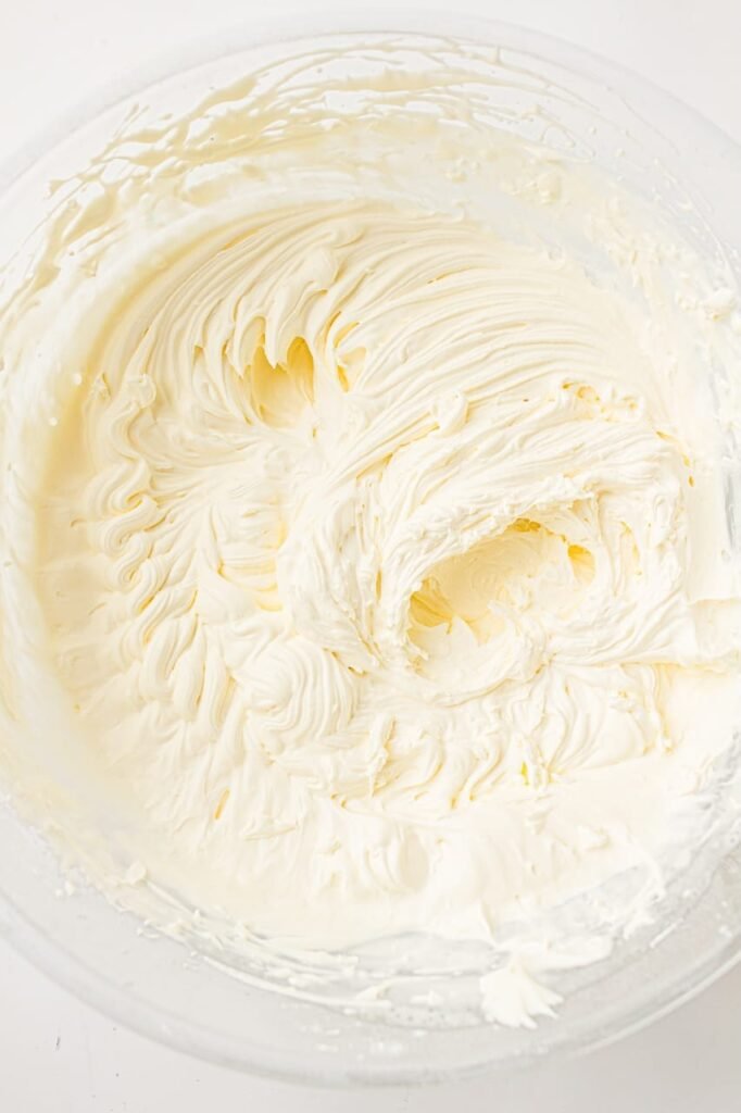 Freshly whipped cream frosting in a clear mixing bowl after being beaten with an electric hand mixer until light and fluffy. The frosting has soft swirls and airy peaks throughout the bowl, showing the finished texture before piping onto homemade red, white, and blue patriotic cupcakes for a 4th of July dessert recipe.