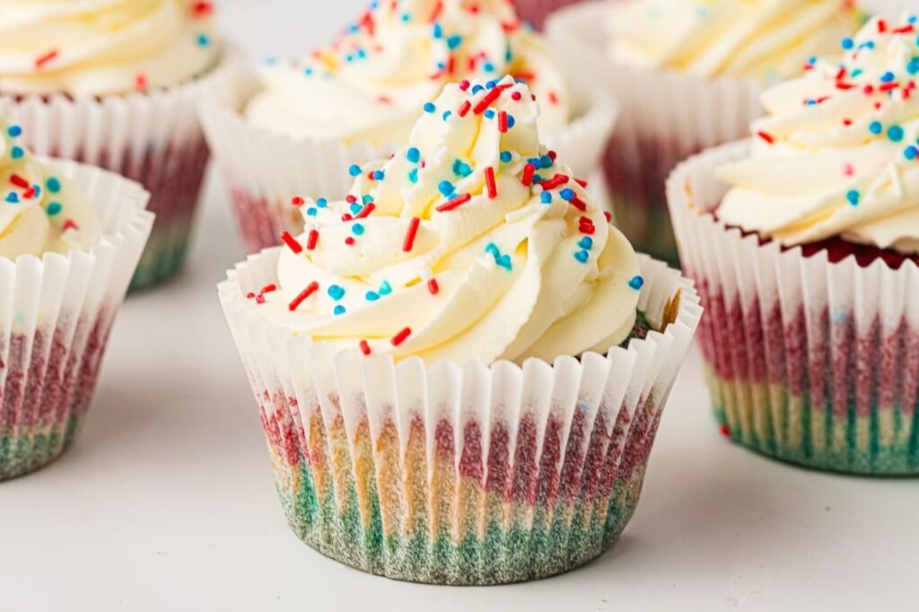 Hero image of patriotic red, white, and blue cupcakes topped with thick swirls of whipped cream frosting and red, white, and blue sprinkles. The front cupcake is sharply focused, showing colorful layered vanilla cake baked inside a white cupcake liner with visible blue, white, and red sections around the edges. Several frosted cupcakes sit softly blurred in the background on a clean white surface, creating a bright and festive summer dessert scene perfect for Fourth of July celebrations, Memorial Day parties, and patriotic gatherings.