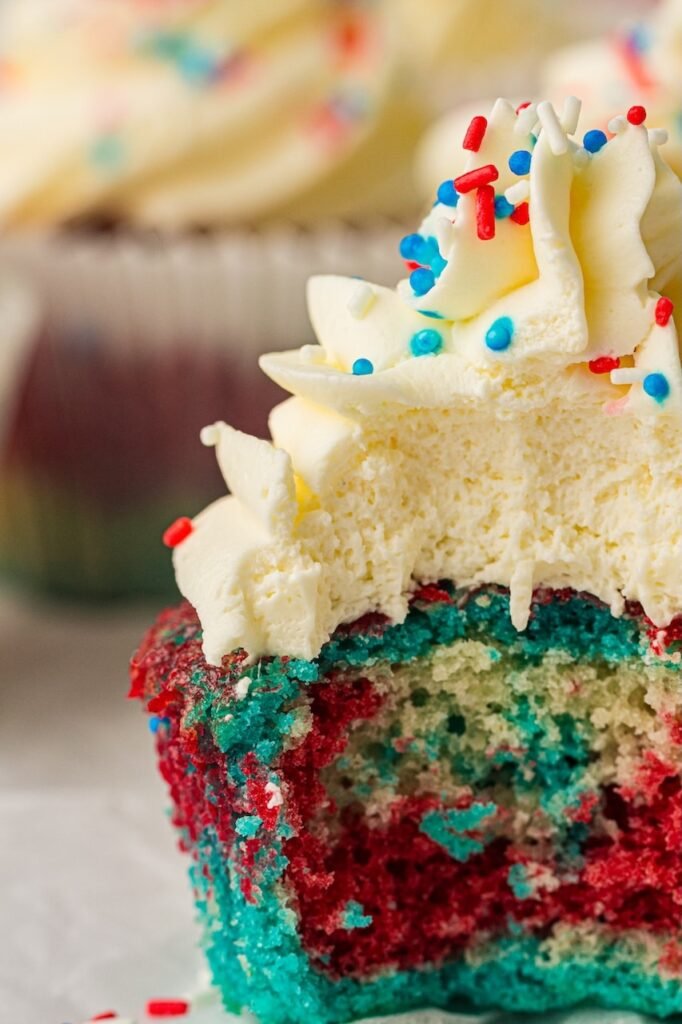 Extreme close-up hero image of a partially eaten patriotic cupcake showing vibrant red, blue, and vanilla cake layers inside a soft, moist crumb. The cupcake is topped with thick swirls of fluffy whipped cream frosting and red, white, and blue sprinkles scattered across the top. The bright blue and red cake colors contrast against the creamy white frosting, while blurred frosted cupcakes sit softly in the background. This detailed interior shot highlights the colorful texture and layered effect of these festive 4th of July cupcakes.