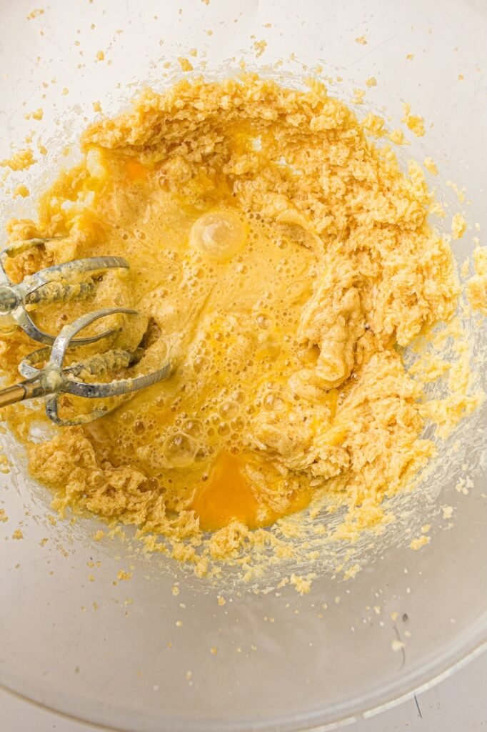Overhead view of eggs and vanilla being added to a creamed butter and sugar mixture in a large glass mixing bowl while making homemade 4th of July M&M cookies. The partially mixed dough appears light and fluffy with visible egg yolks and air bubbles, showing the early stages of creating soft, chewy patriotic cookies from scratch.