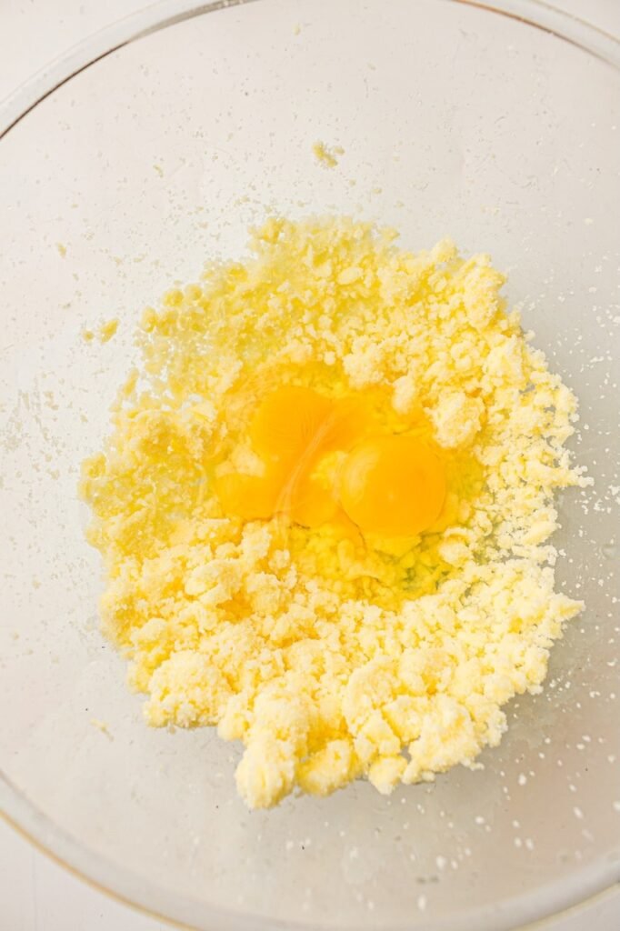 Two eggs sitting on top of a pale yellow butter and sugar mixture in a large clear glass mixing bowl during the early stages of making homemade 4th of July cupcake batter.