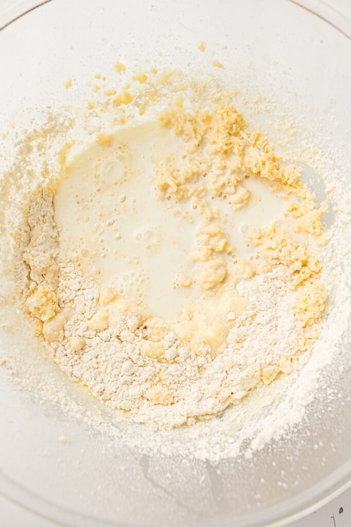 Flour and milk added to a partially mixed butter, sugar, and egg mixture inside a large clear glass bowl while preparing homemade 4th of July cupcake batter. Small clumps of butter mixture are still visible around the edges before the batter is fully combined.