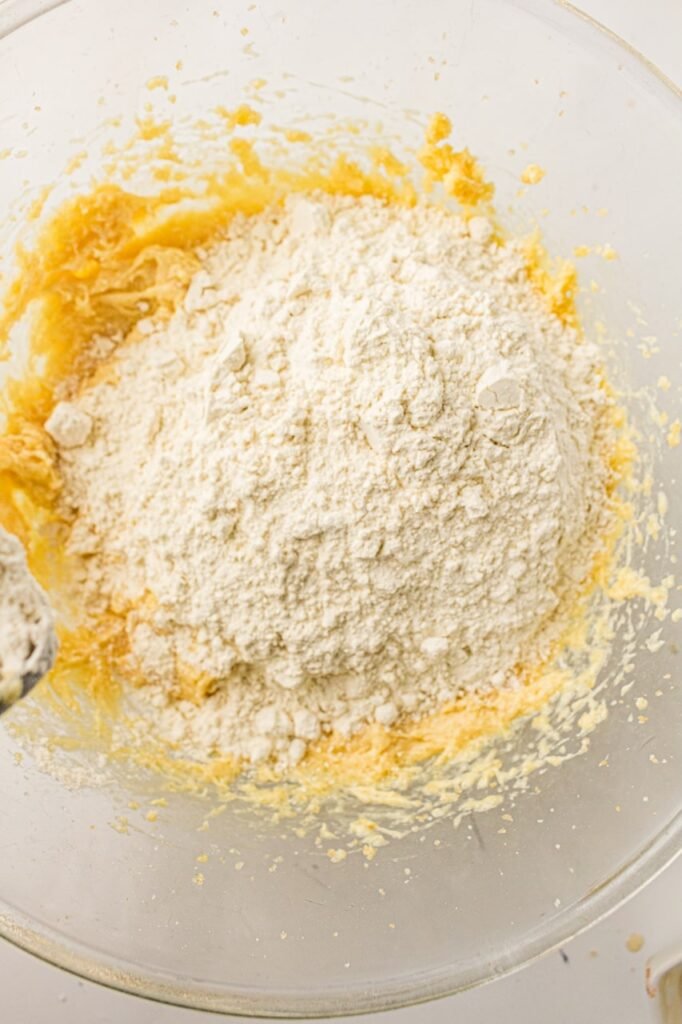 Flour added to a wet cookie dough mixture in a large glass bowl while making homemade 4th of July M&M cookies. The partially mixed dough shows the stage before the dry ingredients are fully combined, helping create soft and chewy patriotic cookies with tender centers and bakery-style texture.