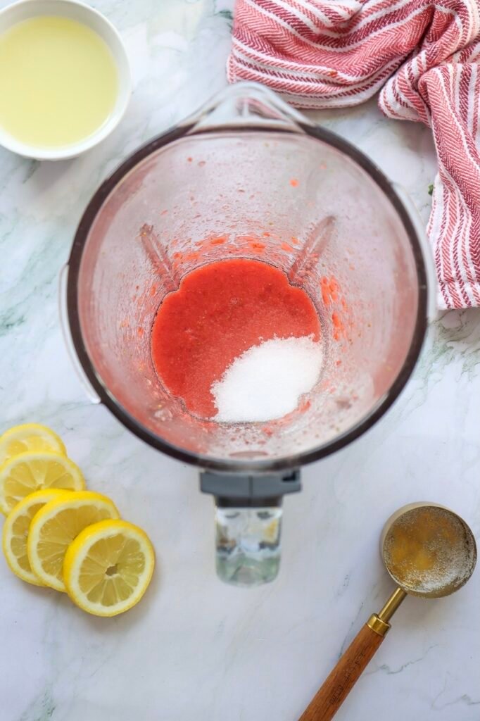 Granulated sugar added to fresh puree inside a blender while making homemade drink with sliced lemons and lemon juice nearby on a marble countertop.