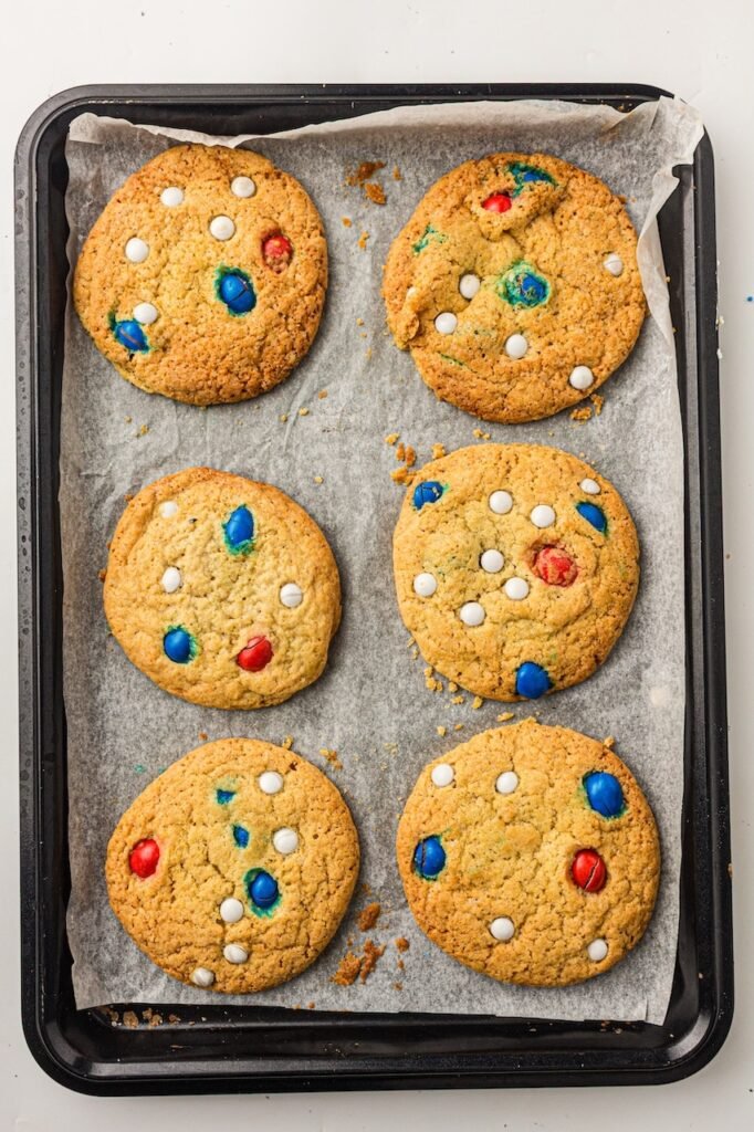 Overhead view of six baked cookies on a parchment-lined baking tray with golden brown edges and red, white, and blue candies baked into the tops. The homemade patriotic cookies appear soft and chewy with thick centers and slightly crisp edges fresh from the oven.