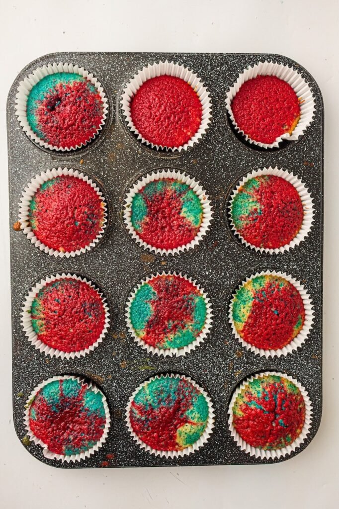 Freshly baked red, white, and blue cupcakes cooling inside white paper liners in a dark speckled muffin pan. The cupcake tops have a colorful marbled patriotic pattern with bright red and turquoise blue sections visible across the domed surfaces after baking.