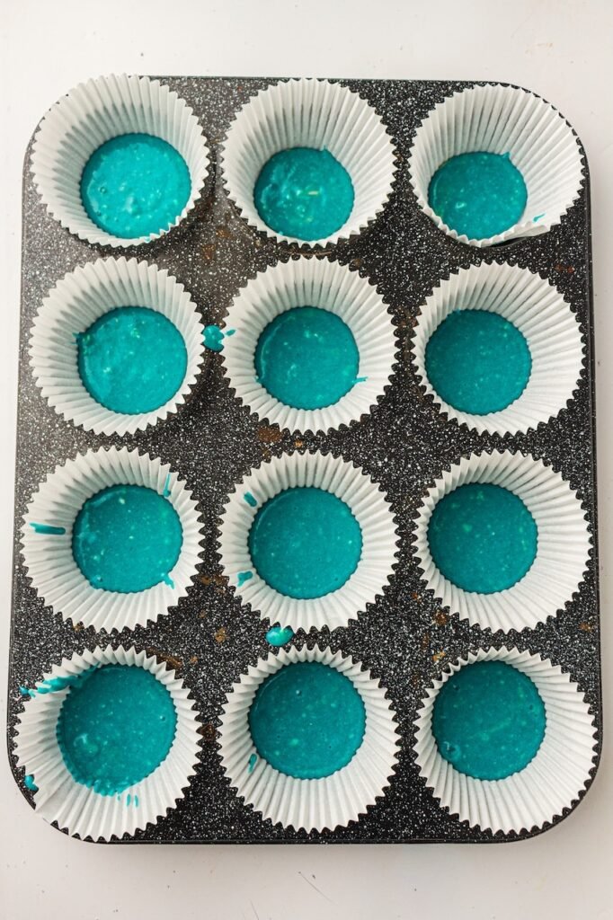 Blue cupcake batter spooned into white paper cupcake liners inside a dark speckled muffin pan while preparing layered 4th of July cupcakes. The bright turquoise-blue batter sits evenly in the bottom of each liner, creating the first patriotic color layer before adding the plain and red batters.