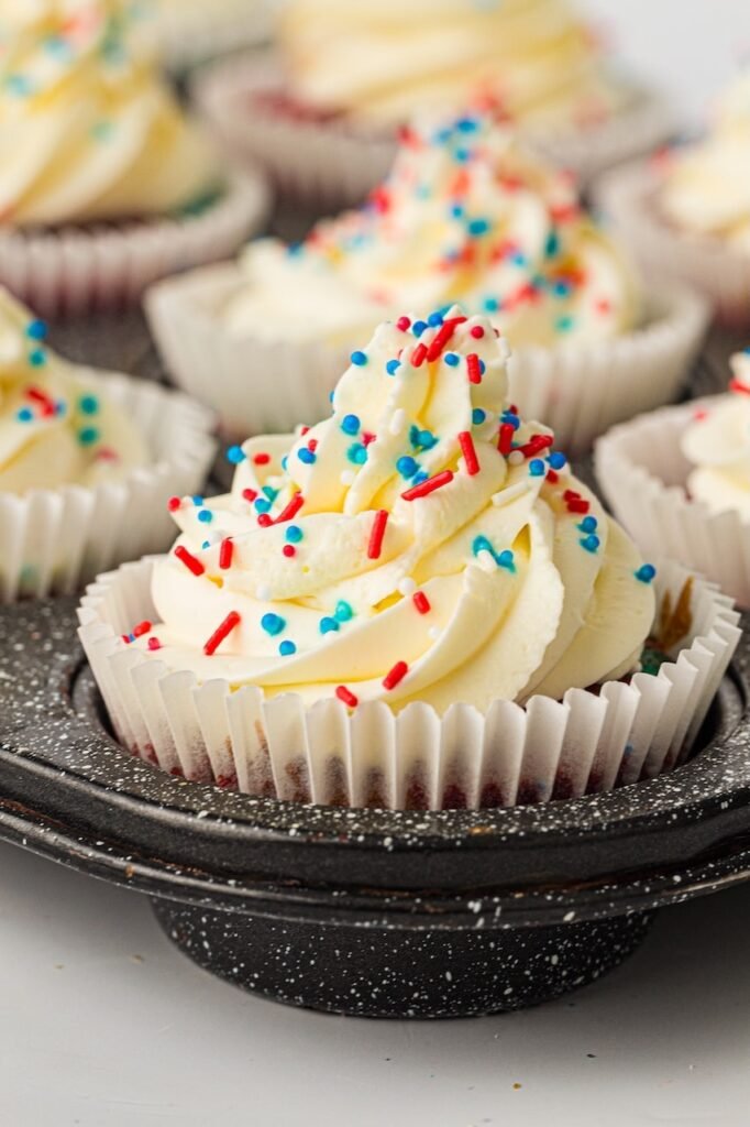 patriotic red, white, and blue cupcakes topped with thick swirls of fluffy whipped cream frosting and colorful red, white, and blue sprinkles. The front cupcake sits inside a dark speckled muffin pan while several frosted cupcakes blur softly in the background. The frosting is piped high with a large star tip, creating dramatic swirls and texture that stand out against the bright sprinkles. The cupcake liners reveal faint patriotic cake colors underneath, making this festive summer dessert perfect for Fourth of July parties, Memorial Day gatherings, and backyard cookouts.