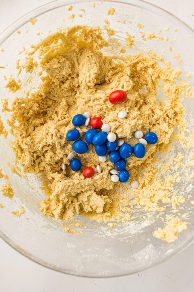 Overhead close-up of thick homemade cookie dough in a glass mixing bowl with red, white, and blue M&Ms being added for festive cookies. The soft buttery dough appears smooth and fluffy while the colorful candies create a patriotic red, white, and blue cookie dough mixture before baking.