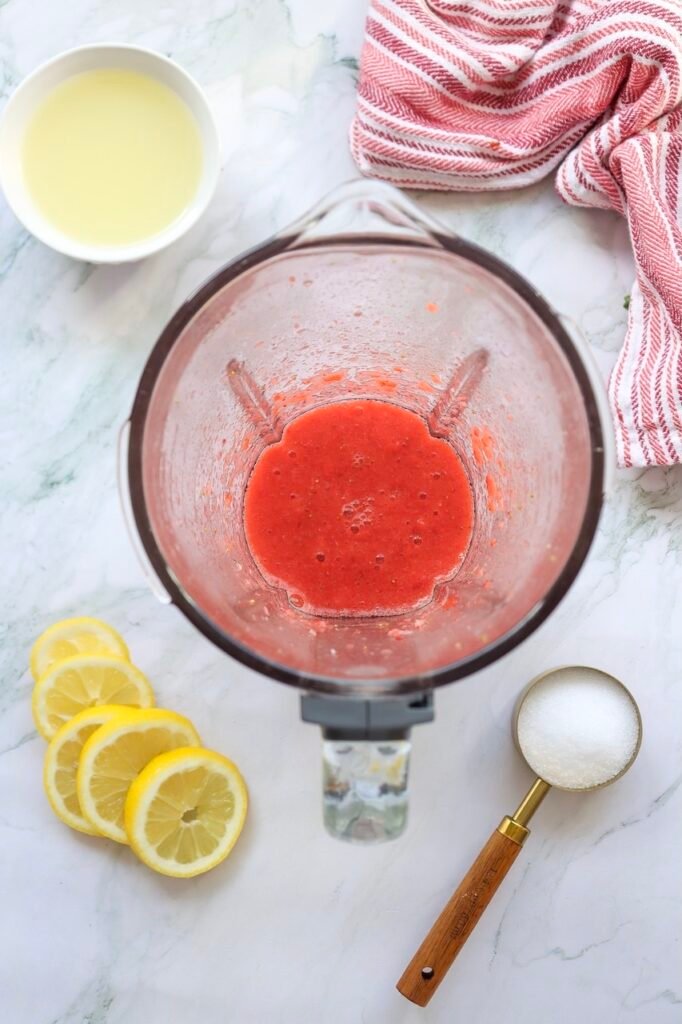 Fresh strawberry puree blended until smooth inside a blender for homemade patriotic strawberry lemonade, surrounded by sliced lemons, sugar, and fresh lemon juice on a white marble countertop.