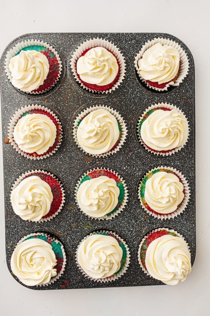 Twelve homemade patriotic cupcakes in white paper liners arranged in a dark speckled muffin pan and topped with thick swirls of freshly piped whipped cream frosting. The cupcakes feature visible red and blue marbled cake layers peeking through the sides, showing the decorated cupcakes before adding festive red, white, and blue sprinkles for a 4th of July dessert recipe.