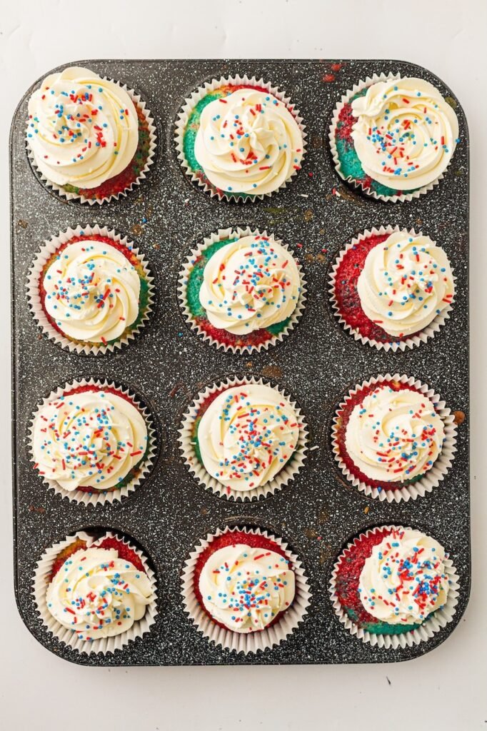 Finished patriotic cupcakes arranged in a dark speckled muffin pan and topped with thick swirls of whipped cream frosting and red, white, and blue sprinkles. The cupcakes have vibrant marbled red and blue cake layers visible around the edges and are decorated for a festive 4th of July dessert presentation.
