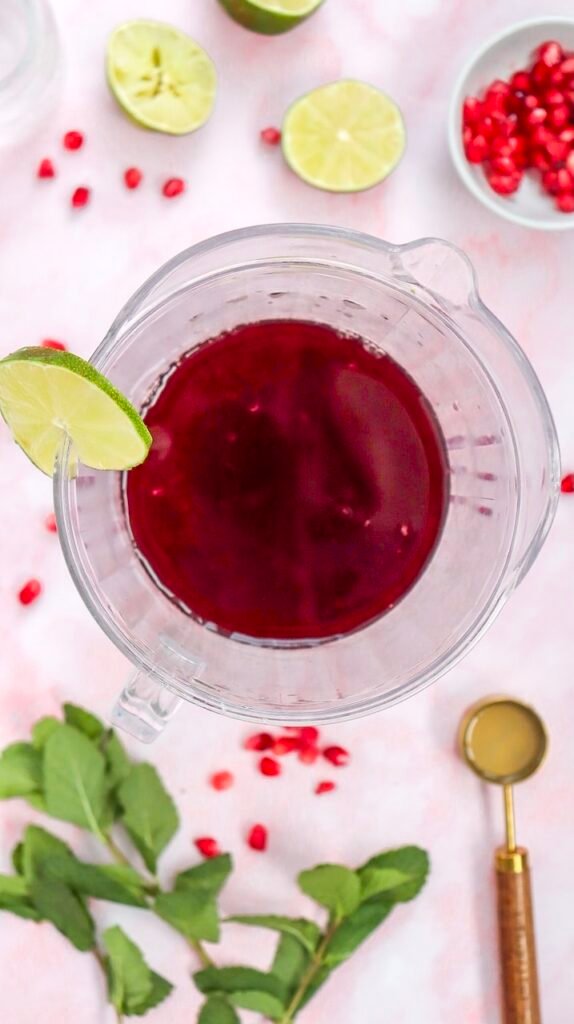 Overhead view of non alcoholic drink mixture in a glass pitcher with lime slices, fresh mint, and pomegranate seeds on a pink marble surface.